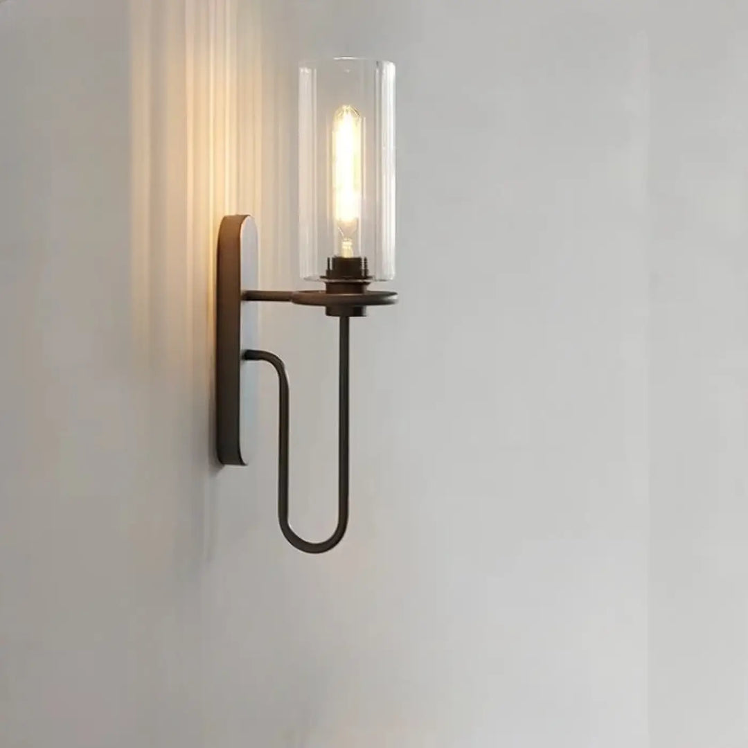 Applique murale loftia en bronze foncé avec bras métallique courbé et cylindre en verre clair abritant une ampoule filament