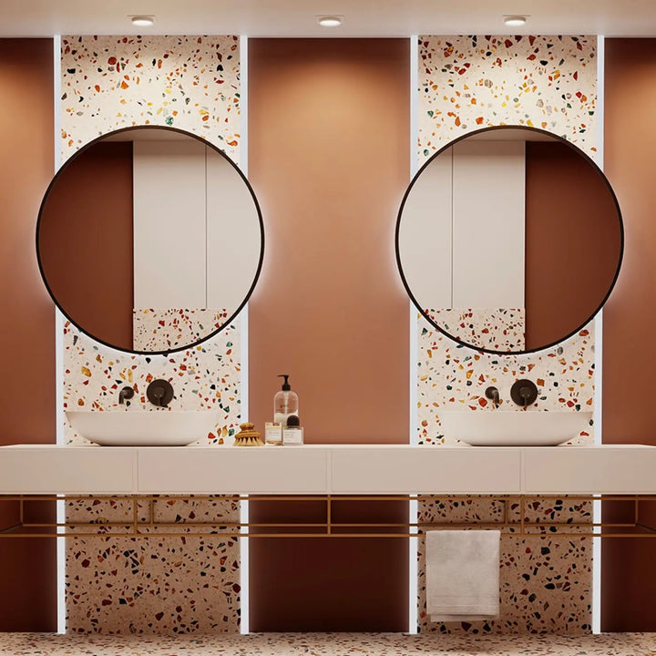 Deux miroirs ronds cadre carré noirs au dessus de deux vasques blanches dans une salle de bain avec murs terrazzo et spot semi-encastré disque lumineux
